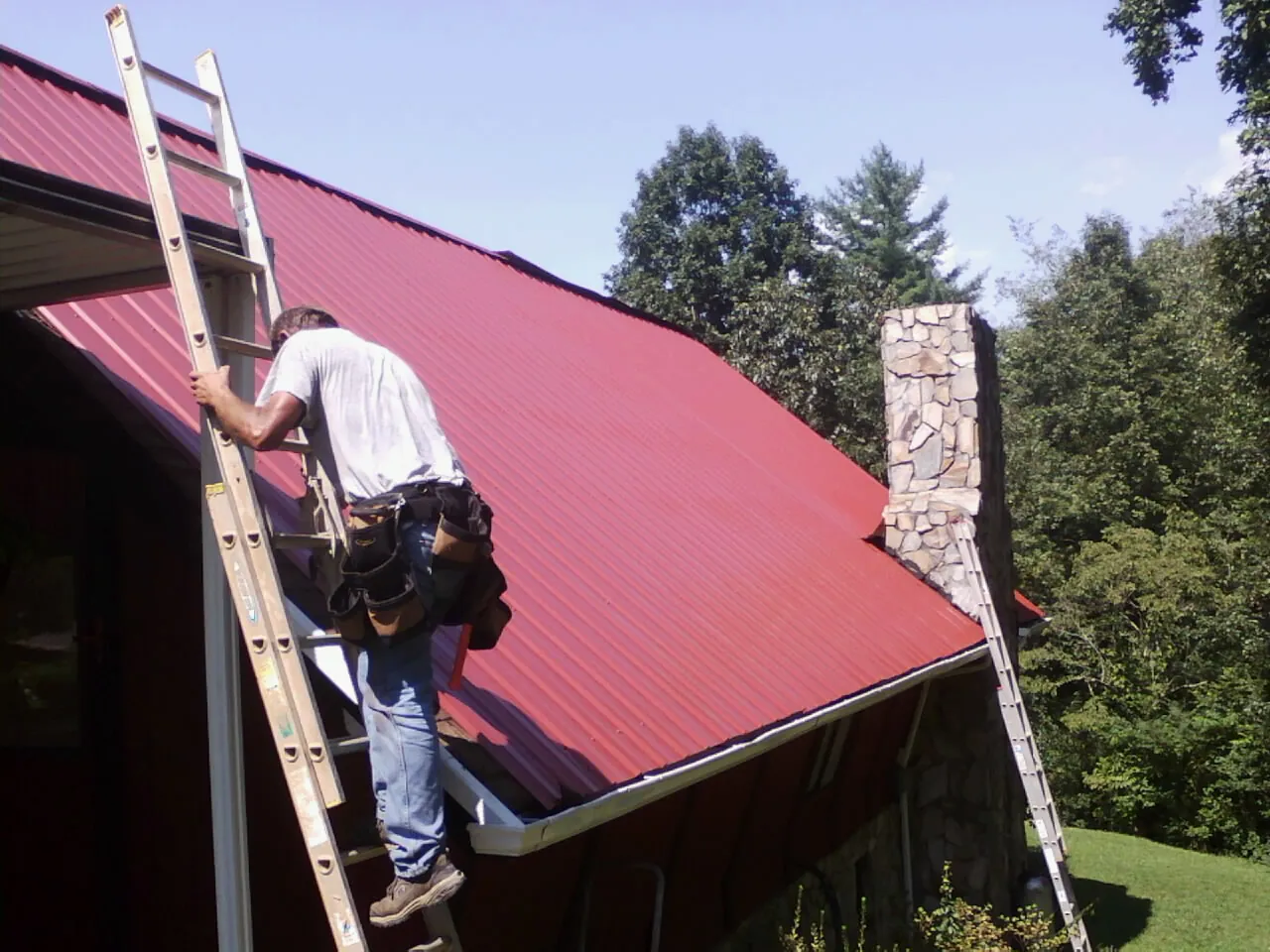 Roofer on a ladder beside a red metal roof and stone chimney — Soffit & Fascia Repair by Roofing Brothers in Troutman